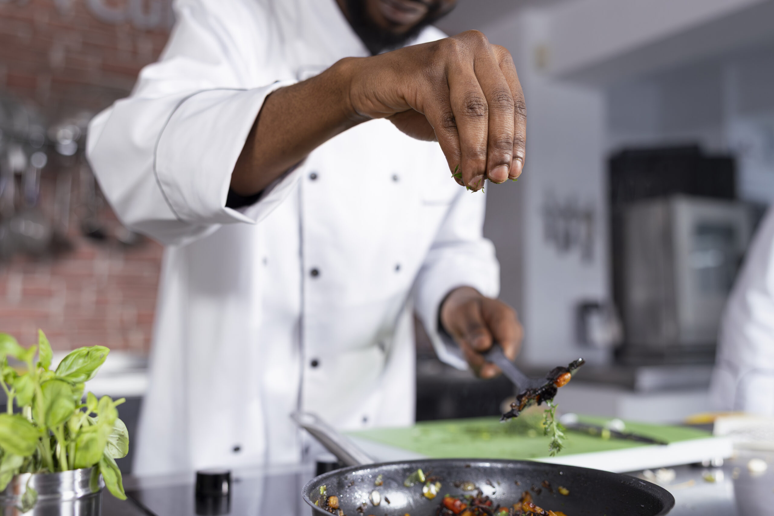 black head chef adding a sprinkle of rosemary to a gourmet dish on the stove